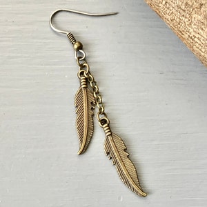 May include: A single earring with two bronze-toned feather pendants hanging from a chain. The earring has a fish hook ear wire. The feathers are detailed with etched lines. The earring is set against a light grey background.