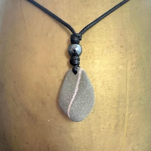 May include: A handcrafted necklace featuring a smooth, gray stone pendant with a pinkish-white stripe. The pendant is suspended from a black cord, accented with a metallic bead. The necklace is a unique, natural accessory.