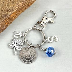 May include: Silver-toned keychain with a clover charm, a coin, the number 70, the letter S, and a blue crystal bead. The coin is dated 1956. The keychain has a lobster clasp.