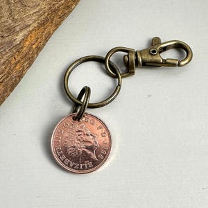 May include: A vintage-style keychain with a 1998 copper coin attached to a bronze-coloured split ring and a lobster clasp. The coin shows a profile of a person and text around the edge. The keychain is set against a neutral background.