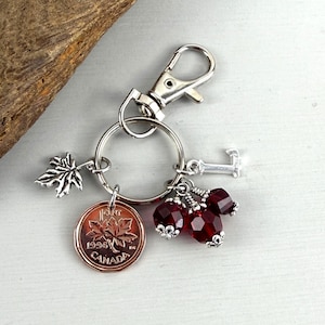 May include: A silver keychain with a lobster clasp, a maple leaf charm, a Canadian penny dated 1996, a letter "L" charm, and three red faceted beads. The penny features a maple leaf design and the word "CANADA".