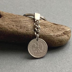 May include: A silver coin keychain featuring a Polish coin with an eagle emblem. The coin is attached to a chain and keyring. The coin is dated 1991. The keychain is displayed on a piece of wood.