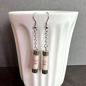 May include: Unusual earrings made from cylindrical components, each displaying "3A" in red. They hang from silver-coloured chain and hook fastenings. The background is a white ceramic container.