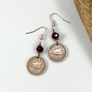 May include: A pair of handcrafted earrings featuring vintage farthing coins from 1947 with a bird design. Each earring has a dark red bead and silver-tone ear wires. The coins are copper-colored.