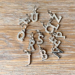 May include: Silver metal alphabet charms with key rings, scattered on a wooden surface. The charms include the letters K, Q, R, S, J, B, A, Z, L, U, and N. Each letter is attached to a small silver key ring.