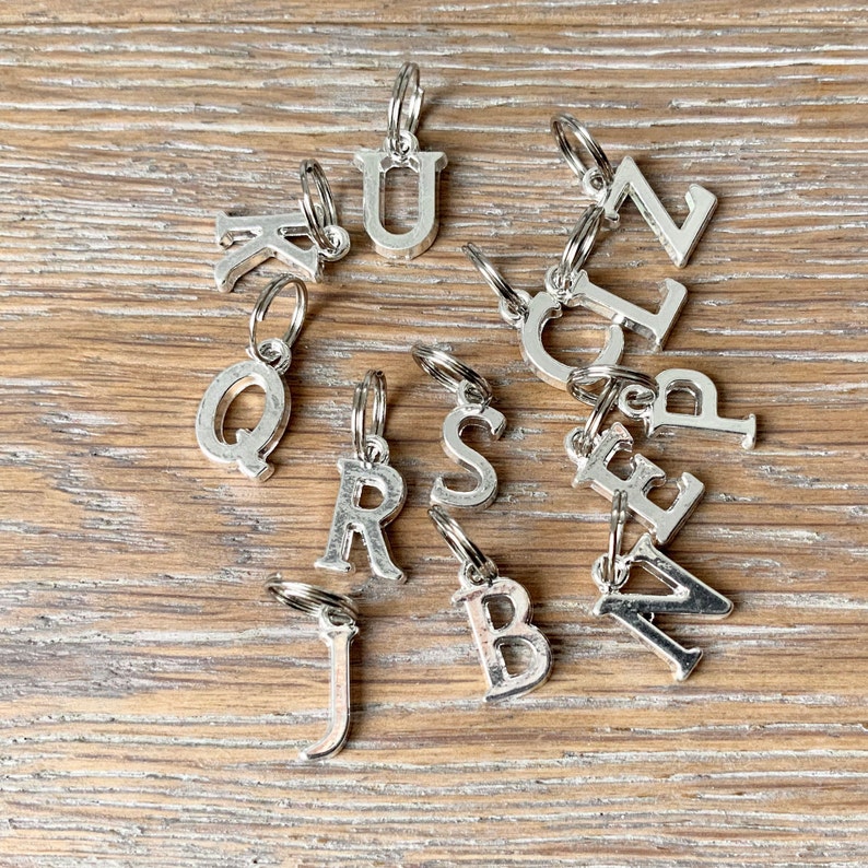 May include: Silver metal alphabet charms with key rings, scattered on a wooden surface. The charms include the letters K, Q, R, S, J, B, A, Z, L, U, and N. Each letter is attached to a small silver key ring.