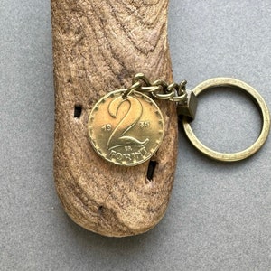 May include: A vintage gold-colored coin keychain. The coin features the number 2, the year 1975, and the word "FORINT". The keychain has a chain and a round key ring. The coin is attached to a piece of wood.