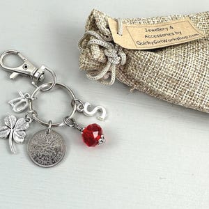 May include: A silver keychain with charms, including a four-leaf clover, a coin, and a red crystal bead. A small burlap bag with a tag that reads "Jewellery & Accessories by QuirkyGirlWorkshop.com" is also present.