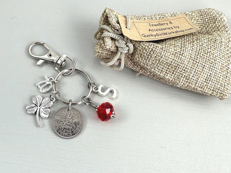 May include: A silver keychain with charms, including a four-leaf clover, a coin, and a red crystal bead. A small burlap bag with a tag that reads "Jewellery & Accessories by QuirkyGirlWorkshop.com" is also present.