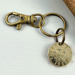 May include: A vintage keychain featuring a brass-toned clasp, ring, and a coin. The coin displays a floral design and the text "THREE PENCE 1939". A unique accessory with a retro feel.