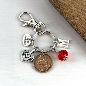 May include: Silver-toned keychain with a lobster clasp, featuring a 'Happy Birthday' charm, a filigree heart, a 1937 farthing coin, a letter 'M' charm, and a red faceted bead. The keychain is designed with multiple charms.
