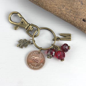 May include: Antique brass keychain with a four-leaf clover charm, a vintage coin, and red faceted beads. The keychain includes a clasp, a large ring, and a letter "M" charm. The coin is dated 1946.