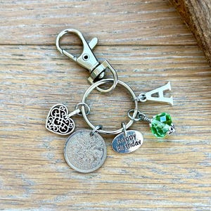 May include: Silver-toned keychain with multiple charms, including a heart, a coin, a green faceted bead, and the letter "A". A small oval charm reads "Happy Birthday". The keychain has a clasp for easy attachment.