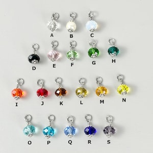 May include: A collection of sixteen colorful crystal charms with silver-tone metal findings. The charms are in various colors, including clear, white, pink, green, orange, red, brown, yellow, blue, and purple. Each charm has a unique shape and sparkle.
