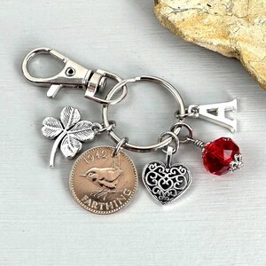 May include: Silver-toned keychain with multiple charms. Charms include a four-leaf clover, a coin with a bird, a filigree heart, a red faceted bead, and a letter "A". The keychain has a lobster clasp.