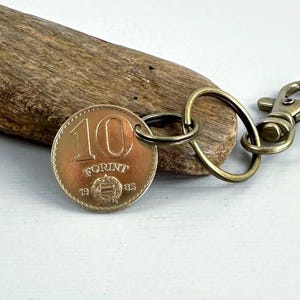 May include: A keychain featuring a bronze-colored 10 Forint coin from 1985. The coin is attached to a brass-colored split ring and a lobster clasp. The coin rests on a piece of driftwood.