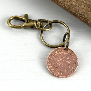 May include: A bronze-colored keychain with a lobster clasp and a round key ring. A copper coin with the profile of a person and the text "ELIZABETH II D.G. REG. F.D. 2012" is attached to the key ring.