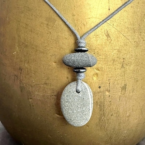 May include: A gray stone pendant necklace with a gray cord. The pendant features an oval stone, a stacked oval stone, and two small, dark gray beads. The necklace is displayed against a gold-colored background.