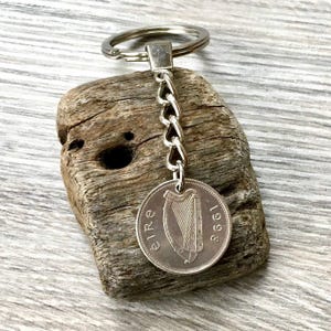 May include: A silver keychain with an Irish coin from 1998, featuring a harp design. The coin is connected to a chain and a key ring. The keychain is displayed on a piece of weathered wood, creating a rustic appearance.