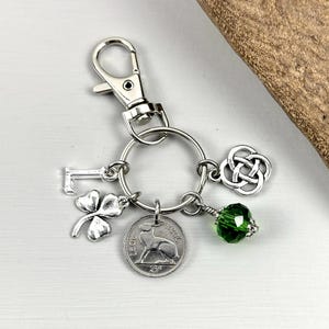 May include: Silver-toned keychain with a lobster clasp, a key charm, a four-leaf clover, a coin with a hare, a Celtic knot, and a green faceted bead. The keychain is on a white surface.