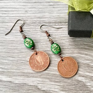 May include: Copper-colored earrings featuring Irish coins dated 2002. Each earring has a green oval bead with a gold shamrock design, connected by copper-toned metal. The earrings are displayed on a wooden surface.