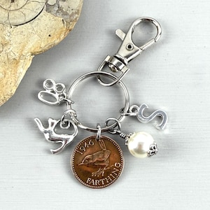 May include: Silver-toned keychain with a lobster clasp, a key ring, and several charms. Charms include a small cat, the number 80, the letter S, a faux pearl, and a Farthing coin from 1946 with a bird design.