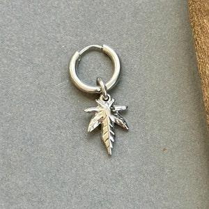 Peut inclure: Boucle d'oreille argentée avec un pendentif en forme de feuille de cannabis. La boucle d'oreille est en métal brillant et présente un charm en forme de feuille détaillé. La boucle d'oreille est présentée sur un fond gris.