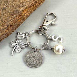 May include: Silver-toned keychain with a clover charm, the number 70, and the letter "S". A vintage coin and a pearl dangle from the key ring. The keychain has a clasp for easy attachment.