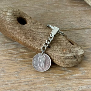 May include: A silver keychain featuring an Irish coin from 1942 with the word "eire" and a harp design. The coin is attached to a chain and a key ring. The background is a piece of driftwood.