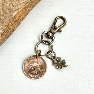May include: A bronze-toned keychain featuring a 1945 farthing coin with a bird design, a four-leaf clover charm, and a clasp. The keychain is designed for attaching to keys or bags.
