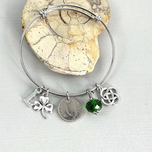 May include: A silver-toned bangle bracelet with charms. Charms include a letter "L", a shamrock, a coin, a green faceted bead, and a Celtic knot. The bracelet is displayed on a light-colored surface.