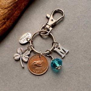 May include: A silver keychain with a clasp, a ring, and several charms. Charms include a four-leaf clover, a coin with a bird, a blue crystal bead, a letter "M", and a charm that says "Happy Birthday".