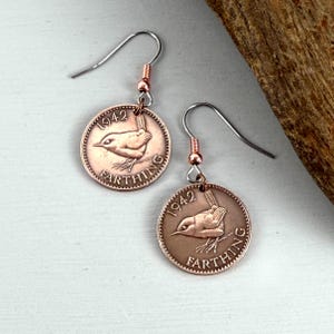 May include: A pair of Farthing coin earrings in a copper colour. Each coin shows a bird design and the year 1942. The earrings have silver-coloured ear wires and copper-coloured accents. The earrings are displayed on a white surface.