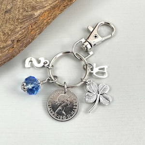 May include: Silver-toned keychain with a lobster clasp, a blue crystal bead, a coin, a four-leaf clover charm, and the number "2". The coin features a profile of a person. The keychain is attached to a wooden surface.