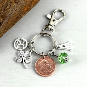 May include: Silver keyring with a lobster clasp and multiple charms. Charms include a four-leaf clover, a coin, the number 20, a letter "A", and a green crystal bead. The coin features a profile of a person.