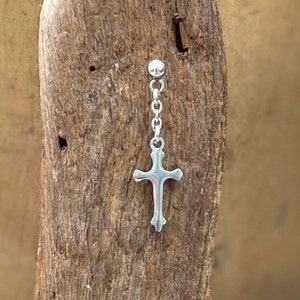 May include: A silver earring with a cross pendant hanging from a chain. The cross is detailed and suspended from a post with a small ball. The earring is set against a textured wooden background.