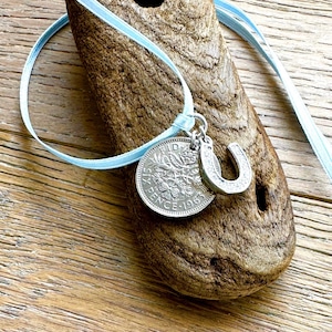 May include: A silver pendant necklace with a vintage coin and a horseshoe charm, suspended from a pale blue ribbon. The coin is dated 1963. The necklace is displayed on a piece of driftwood.