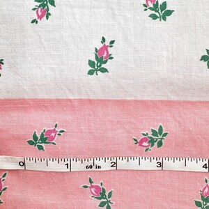 May include: Close-up of a fabric with a repeating floral pattern. The design features pink rosebuds with green leaves on a white and pink background. A measuring tape is placed across the fabric, showing inches.