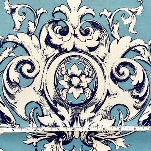 May include: Light blue fabric with a repeating white and navy blue baroque design. A measuring tape shows the fabric's width. The fabric features a floral motif within a circular frame, surrounded by ornate scrollwork.