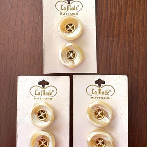 May include: Three sets of La Mode Buttons on their original cards. Each card holds two pearlescent, round buttons with a gold-colored cross design in the center. The cards are cream-colored with the brand name printed in a decorative font.