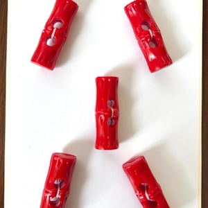 May include: A collection of five bright red toggle buttons with a bamboo-like design. Each button has two holes for sewing and a textured surface. The buttons are arranged on a white background.