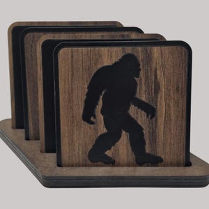 May include: Set of four brown wooden coasters with a black silhouette of Bigfoot walking. The coasters are in a holder made of the same wood.