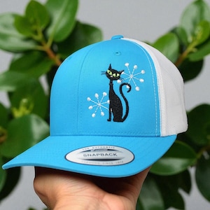 May include: A blue and white trucker hat featuring a black cat design with yellow eyes and white accents. The cat is embroidered on the front of the hat, which has a curved brim and a mesh back.