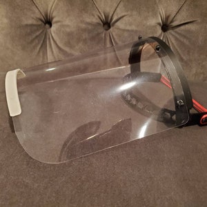 May include: A clear plastic face shield with a black headband and a white adjustable strap. The shield is designed to protect the wearer's face from splashes and droplets.