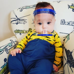 May include: A baby wearing a blue and yellow shirt with a clear plastic face shield. The shield has a blue headband. The baby is sitting in a chair with a white and green patterned fabric.