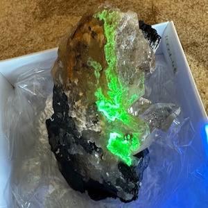 May include: A large, clear quartz crystal with a green fluorescent mineral embedded inside. The crystal is sitting on a dark gray rock.