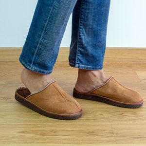 May include: A pair of brown suede slippers with a white fur lining. The slippers are worn by a person wearing blue jeans.