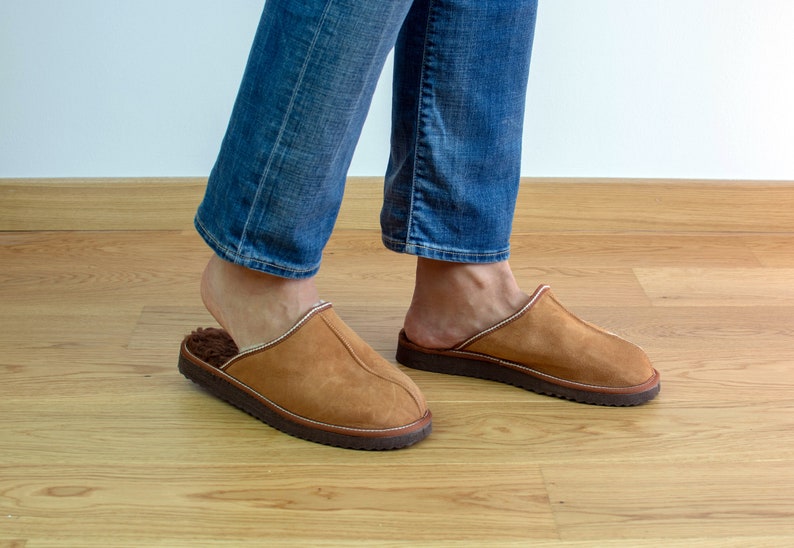 May include: A pair of brown suede slippers with a white fur lining. The slippers are worn by a person wearing blue jeans.