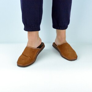 May include: A pair of brown suede slippers with a closed toe and an open back. The slippers have a brown leather trim around the edges and a brown leather sole.