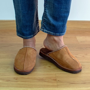 May include: A pair of brown suede slippers with a white trim. The slippers are worn on a wooden floor.